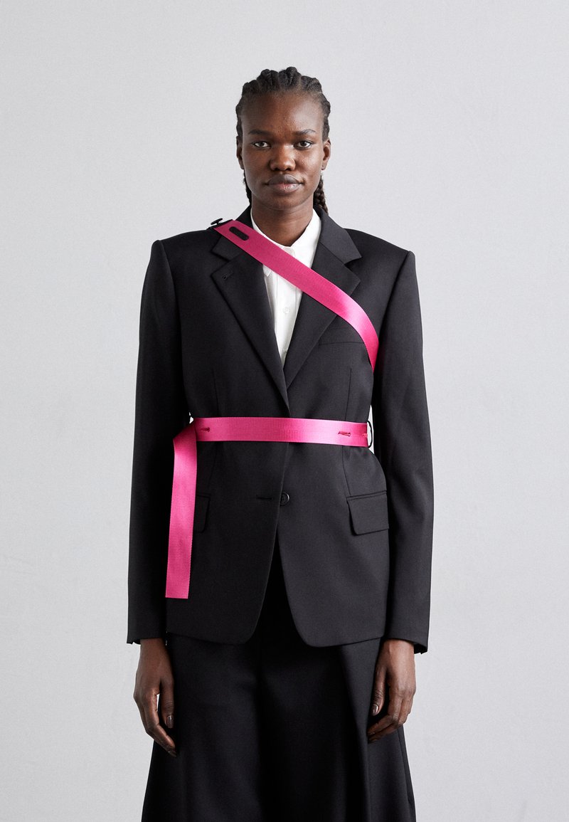 Blazer noir ajusté avec des revers nets, associé à une chemise blanche. Une ceinture rose vif est drapée sur le torse et l'épaule, ajoutant un contraste.