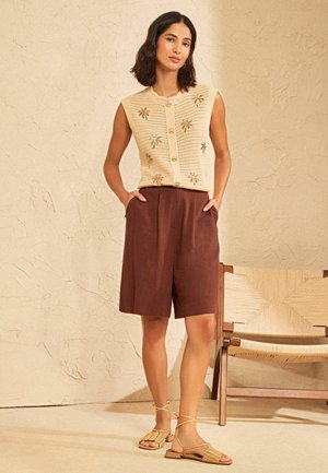 Kvinde iført ærmeløs beige strikket top med blomsterbroderi, brune højtaljede shorts og beige sandaler, stående ved siden af en flettet stol.