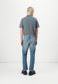 Camisa polo gris, jeans rectos de mezclilla azul claro, zapatos gruesos negros. La mezclilla cuenta con bolsillos y una etiqueta de cuero en la parte trasera de la cintura.