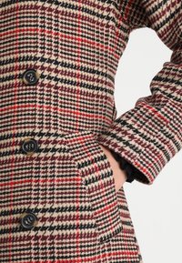 Cappotto in misto lana con motivo a carlino in beige, rosso e nero. Presenta bottoni neri e un elegante dettaglio a balza sui polsini.