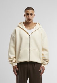 Cremfarbener Reißverschluss-Hoodie mit großzügigem Schnitt, großer Kapuze und Fronttaschen; getragen über einem weißen Shirt und braunen Hosen.
