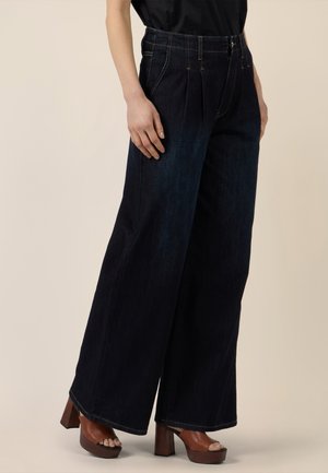 Persona vistiendo jeans oscuros de pierna ancha con pliegues, una blusa negra y tacones de plataforma marrones con punta abierta, de pie frente a un fondo beige.