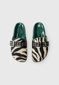 Musta ja kerma zebra-kuvioinen liukuva klompi mustalla hihnalla, jossa on hopeanvärisiä niittejä, vihreät sisäpohjat ja pyöreä kärki.