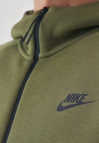 Primer plano de una sudadera con cremallera de color verde oliva de Nike, con cremallera negra y el logo negro de Nike en el pecho.