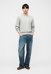 Νέος άνδρας με μαύρα μαλλιά, ντυμένος με ανοιχτό γκρι φούτερ, μπλε τζιν παντελόνι με φαρδιά πόδια, καφέ αθλητικά παπούτσια, στέκεται με τα χέρια στις τσέπες.