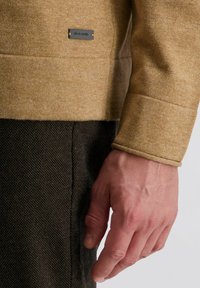 Pull en texture marron avec une teinte dorée, arborant une étiquette de marque. Porté sur un pantalon à motifs sombres, mettant en avant une coupe décontractée et des manches retroussées.