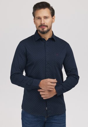 Mężczyzna z przyciętą brodą, ubrany w granatową koszulę w kropki z długim rękawem i niebieskie dżinsy, zapinający mankiet z rękami splecionymi przed sobą.