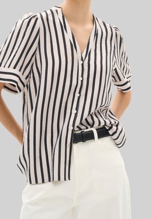 Person iført en beige kortærmet bluse med lodrette sorte striber og hvide knapper, kombineret med hvide bukser og et sort bælte.