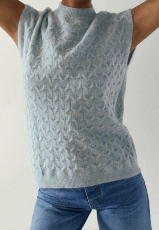 Pull sans manches gris clair en maille avec un motif torsadé porté avec un jeans en denim bleu, bras levés au-dessus de la tête.