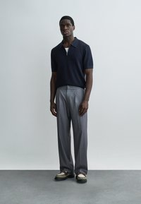 Fiatal férfi áll egy szürke fal előtt, sötétkék rövid ujjú pólót, szürke redős nadrágot és fehér, sötét talpú mokaszint visel.