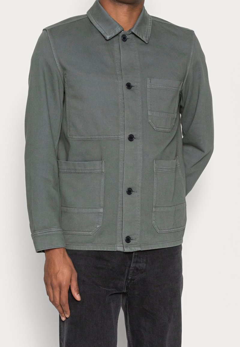 Homme portant une veste grise boutonnée avec trois poches avant et un pantalon noir, debout avec une main le long du corps.