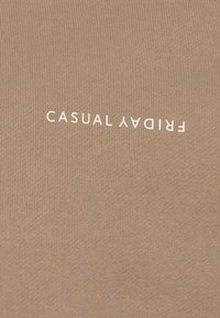 Lys brun stoff med ribbet tekstur som har teksten "CASUAL FRIDAY" i hvitt, vist horisontalt over midten.