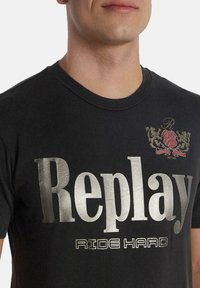 Camiseta de algodón negra con texto metálico en plata "Replay" y "RIDE HARD" debajo. Incluye un pequeño logo bordado en rojo y dorado.