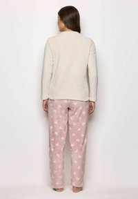 Donna in piedi a piedi nudi, indossa un top in pile lungo a maniche cremisi e pantaloni in pile rosa con motivi a cuori bianchi, di spalle rispetto alla fotocamera.
