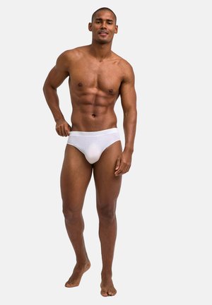 Mężczyzna o umięśnionej sylwetce stojący boso, ubrany w białe bokserki, lekko naciągający gumkę, na jasnym, jednolitym tle.