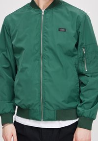 Grønn nylon bomberjakke med ribbestrikket krage og mansjetter. Frontglidelås, glidelåslomme på venstre erme, og glatt tekstur. Justerbar passform.