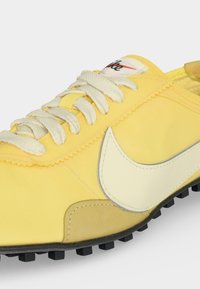 Κίτρινο αθλητικό παπούτσι Nike με λευκό λογότυπο swoosh, λευκά κορδόνια και μαύρη υφασμάτινη σόλα σε κοντινή λήψη.