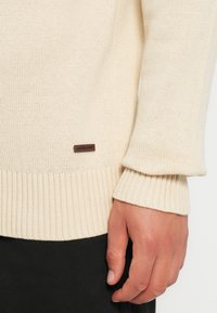 Lys beige strikkegenser med ribbede mansjetter og nederkant. Har en liten brun etikett med tekst nær bunnen. Myk tekstur, lett design.