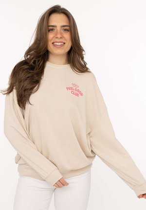 Smiling woman with long brown hair wearing a beige sweatshirt with pink "Feel Good Club" text and white pants on a white background.