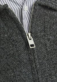 Graue Strickjacke mit Reißverschluss aus strukturiertem Material, silbernem Reißverschluss und einem gestreiften Hemd, das darunter sichtbar ist.