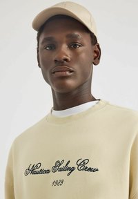 Sudadera beige con bordado negro "Nautica Sailing Crew" y acento "1983", combinada con una camisa blanca. Gorra beige completa el conjunto.