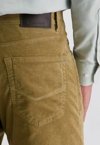Pantalons en velours côtelé marron clair avec une poche arrière et une étiquette de marque en cuir marron foncé, associés à une chemise à manches longues texturée de couleur claire.