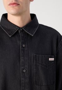 Black denim shirt with a button-down collar, front pocket and red Calvin Klein logo tag, featuring a textured finish and subtle stitching details.