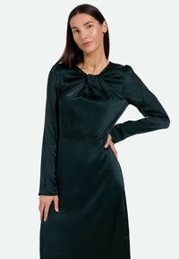 Vestido de manga larga en satén verde oscuro, que presenta un detalle anudado en el escote y una textura suave, con una silueta de longitud media.