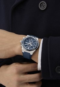 Sudraba un zilā krāsā hronogrāfa pulkstenis ar teksturētu zilo ciparnīcu, gumijas siksniņu un nerūsējošā tērauda akcentiem, kas uzvilkts uz plaukstas.