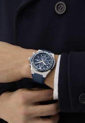 Reloj cronógrafo plateado y azul con una esfera azul texturizada, correa de goma y detalles en acero inoxidable, usado en una muñeca.