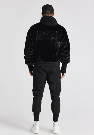 Mężczyzna w czarnej futrzanej kurtce z logo "SIKSILK", czarnych spodniach typu cargo, białych trampkach i czarnej czapce, stojący tyłem na białym tle.
