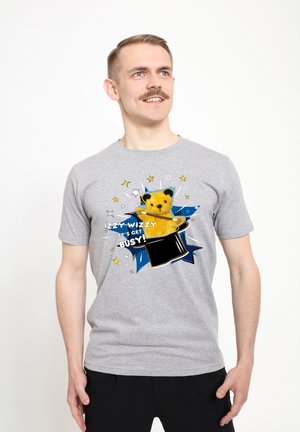 Šedé bavlněné tričko s grafikou žlutého medvěda, který vystupuje z černého klobouku kouzelníka, s modrými hvězdicovými akcenty a textem.