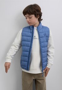 Chaleco acolchado de color azul claro con cuello alto, utilizado sobre una sudadera blanca. Cierre delantero con cremallera y detalle de logo en el pecho.