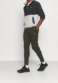 Fleece jas met rits in grijs en zwart met geel logo; groene taps toelopende joggers; zwarte sportschoenen; witte sokken.