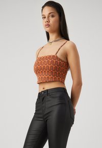 Top corto arancione con motivo ondulato, dotato di spalline sottili. Abbinato a pantaloni neri aderenti, che aggiungono contrasto all'outfit.