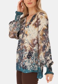 Wielokolorowa bluzka w kwiaty z głębokim dekoltem w kształcie litery V, lekkimi rękawami i turkusowymi marszczonymi mankietami. Charakteryzuje się mieszanką brązowych, niebieskich i kremowych odcieni.