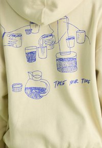 Sweatshirt amarelo com desenhos lineares azuis de várias canecas e um jarro, com o texto "TOMA O TEU TEMPO" integrado no design.
