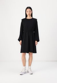 Black dress with long sleeves, lace trim at shoulders, cinched waist, and flared skirt. Worn with white high-top sneakers.
