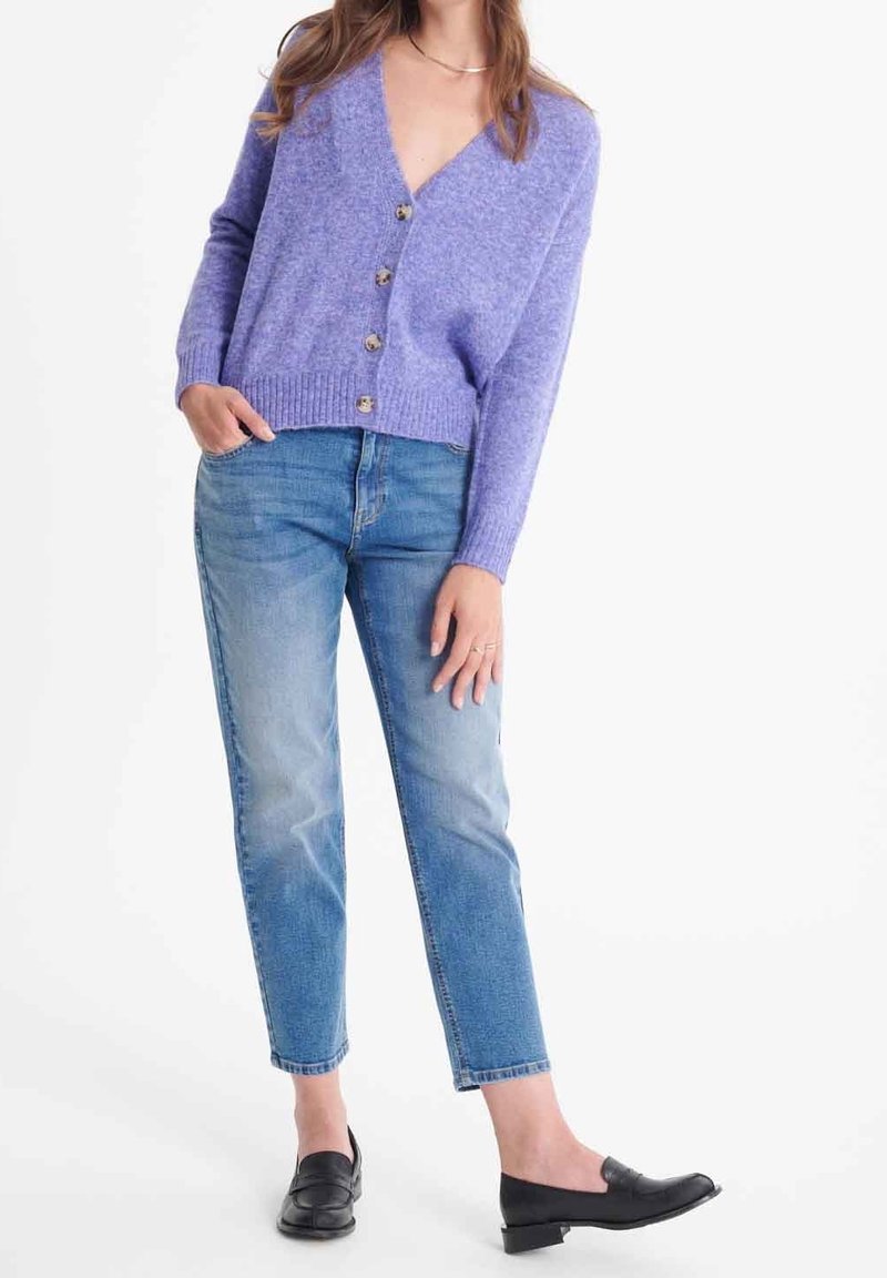Femme portant un cardigan boutonné lavande, un jean bleu à jambes droites, des mocassins noirs, une main dans la poche et l'autre détendue.