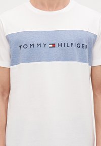 Bijela majica s plavom horizontalnom prugom preko prsa. Sadrži "TOMMY HILFIGER" logo u crnoj boji s crvenim naglaskom. Kratki rukavi.