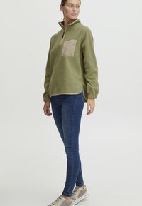 Pullover in pile verde oliva con tasca sul petto beige, design a mezza zip e polsini elasticizzati, abbinato a jeans skinny scuri e scarpe da ginnastica chiare.