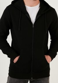 Felpa nera con zip, realizzata in tessuto strutturato, con tasche a marsupio e cappuccio regolabile con coulisse. I polsini e l'orlo ribattuti offrono un look aderente.