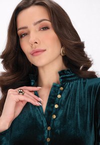 Bluza z aksamitu w kolorze teal z falbaniastym kołnierzem, złotymi guzikami z przodu i ręką ozdobioną złotym pierścionkiem. Gładka tekstura.