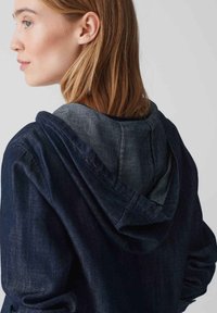 Giacca di jeans con una finitura blu scuro e un cappuccio blu chiaro. Presenta una vestibilità comoda, cuciture prominenti e un cordoncino regolabile per il cappuccio.