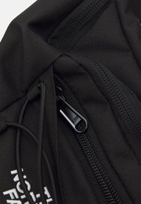 Musta nailonireppu, jossa on tekstuuriinen kangas, vetoketju mustalla metallivetimellä ja valkoinen logo brodeerattuna etupuolelle.