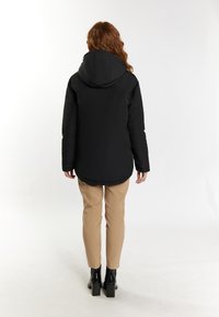 Chaqueta negra acolchada con capucha, con un dobladillo curvado y bolsillos laterales, llevada sobre pantalones beige y botas de tacón negras.