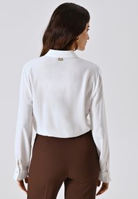 Blusa bianca a maniche lunghe con polsini abbottonati, dotata di colletto e accento del logo dorato sul retro, abbinata a pantaloni marroni.