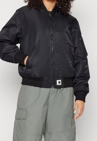 Zwarte bomberjack van gladde stof, met een ritssluiting aan de voorkant, geribde manchetten en een logo patch op de onderste zoom.