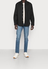 Camicetta nera a bottoni sopra una maglietta chiara, jeans blu e sneakers bianche con accenti arancioni. Presenta una tasca sul petto e una vestibilità comoda.