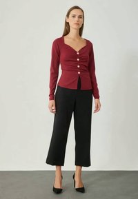 Burgundi színű, testhezálló kardigán mély dekoltázzsal, redős elejével és gombos részleteivel, fekete, bő szárú nadrággal és fekete magassarkú cipővel párosítva.